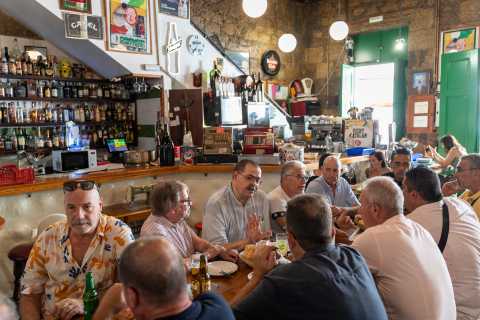 Vista general de la mesa del almuerzo, mientras se ven varias conversaciones simultáneas. De cara, de izquierda a derecha, se ve a Manzano, Jesús Pinar, Saúco, Luis, Julio y Óscar. De espaldas, también de izquierda a derecha, se ve a Diego, Terrón, Eloy, Esteban y a Juan Ramón.